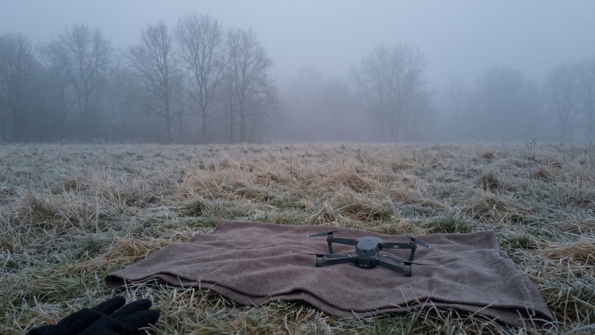 Campo gelato all'alba con la nebbia che nasconde gli alberi e un piccolo drone appoggiato su una coperta sull'erba brinata