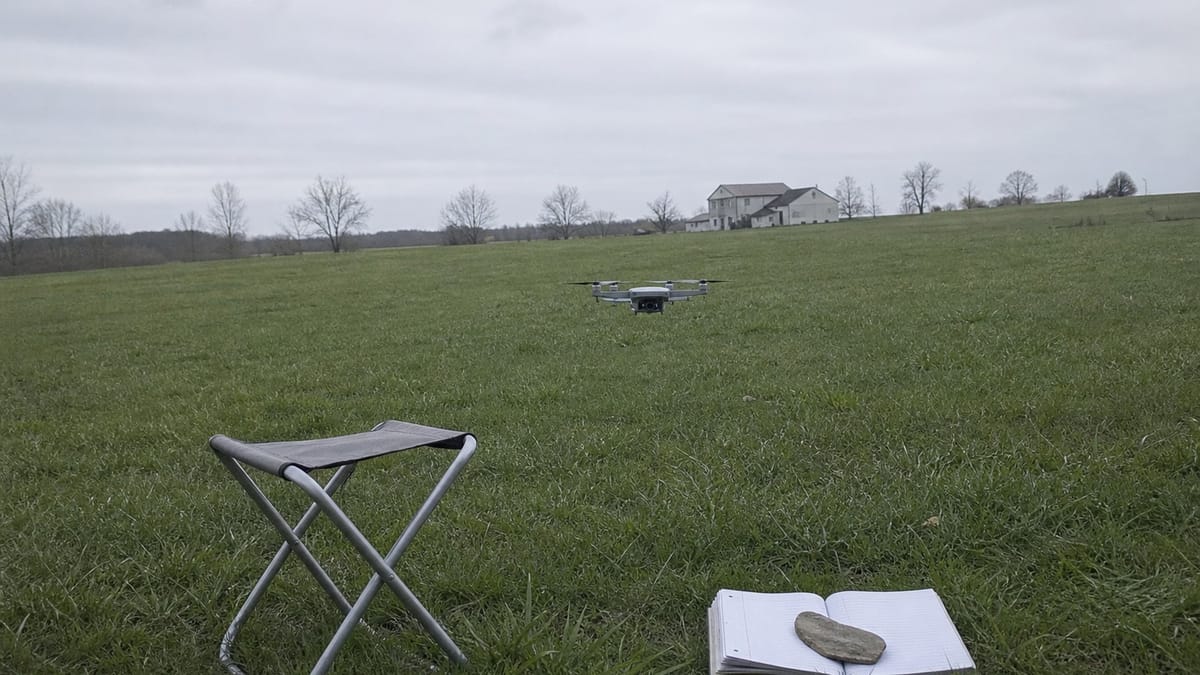 Campo arato e vuoto a inizio primavera con un piccolo drone in volo basso, uno sgabello pieghevole e un quaderno aperto in primo piano