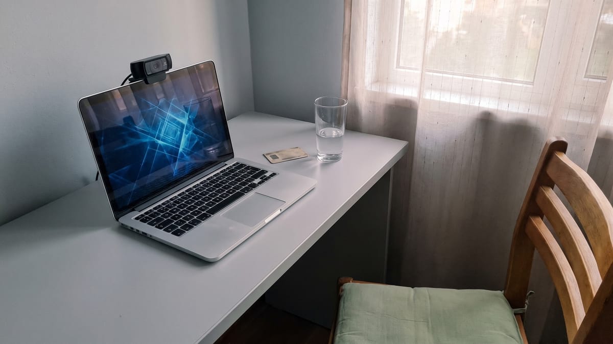 Angolo studio di casa con un portatile aperto, una webcam sopra lo schermo, un documento capovolto e un bicchiere d'acqua