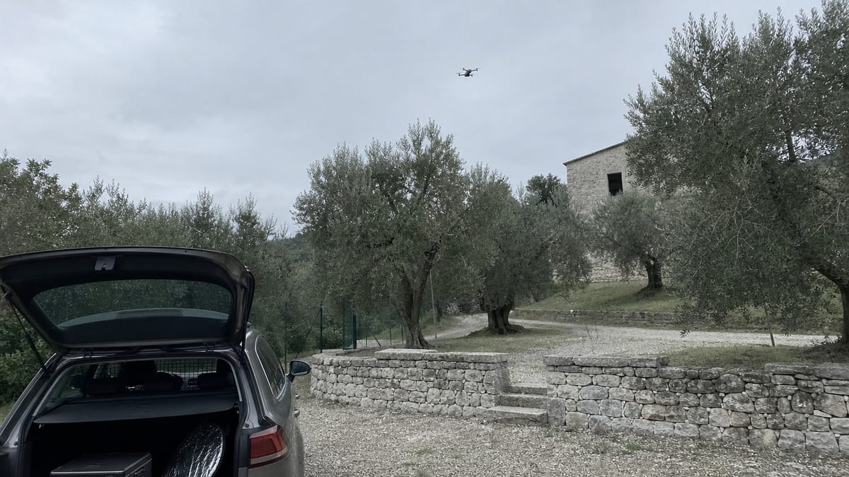 Cortile di un casale di campagna al tramonto con un drone piccolo in volo e il bagagliaio aperto di un'auto in primo piano